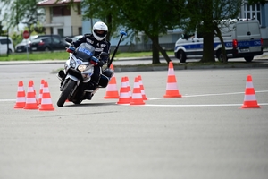 Policjanci na placu manewrowym - podczas konkurencji jazdy motocyklem. Policjant w kasku i na motocyklu jedzie po torze.