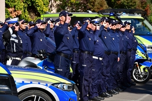Policjanci oddają honory zmarłemu Koledze - upamiętnienie pod budynkiem Komendy Wojewódzkiej Policji w Rzeszowie, po prawej i lewej stronie stoją policyjne radiowozy z włączonymi sygnałami świetlnymi i dźwiękowymi.