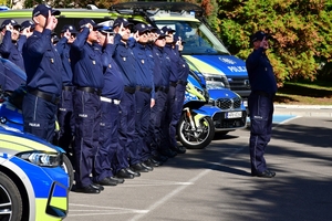 Policjanci oddają honory zmarłemu Koledze - upamiętnienie pod budynkiem Komendy Wojewódzkiej Policji w Rzeszowie, po prawej i lewej stronie stoją policyjne radiowozy z włączonymi sygnałami świetlnymi i dźwiękowymi.