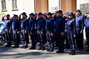 Policjanci oddają honory zmarłemu Koledze - upamiętnienie pod budynkiem Komendy Wojewódzkiej Policji w Rzeszowie, po prawej i lewej stronie stoją policyjne radiowozy z włączonymi sygnałami świetlnymi i dźwiękowymi.