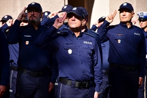 Policjanci oddają honory zmarłemu Koledze - upamiętnienie pod budynkiem Komendy Wojewódzkiej Policji w Rzeszowie, po prawej i lewej stronie stoją policyjne radiowozy z włączonymi sygnałami świetlnymi i dźwiękowymi.