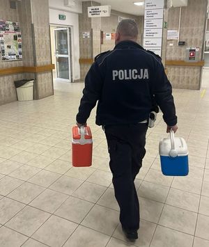 Na zdjęciu od tyłu policjant ruchu drogowego,  na szpitalnym korytarzu. W prawej ręce niesie pojemnik koloru niebieskiego, w lewej czerwonego.