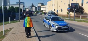 Na zdjęciu policyjny radiowóz, z lewej strony idąca tyłem osoba owinięta tęczową flaga, w tle uczestnicy Marszu Równości.