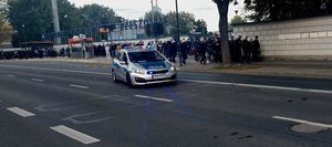 Na zdjęciu policyjny radiowóz i umundurowani policjanci którzy prowadzą kibiców na stadion.