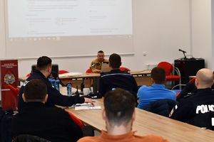 Na zdjęciu umundurowani policjanci  siedzą przy stołach konferencyjnych, na środku sali stoi strażak.  W tyle po lewej stronie tablica  z logo i napisem Państwowa Starz Pożarna.