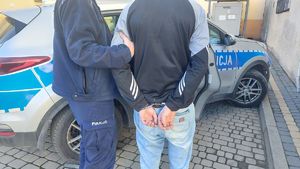 Na zdjęciu umundurowany policjant przytrzymuje mężczyznę, który na rękach trzymanych z tyłu ma założone kajdanki. Obaj stoją przed policyjnym radiowozem.