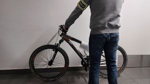 Na zdjęciu policjant w ubraniu cywilnym, na lewym przedramieniu żółta opaska z napisem policja. Policjant prowadzi rower koloru czarnego.