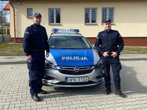 Na zdjęciu dwóch umundurowanych policjantów  na tle radiowozu policyjnego. W tle budynek.