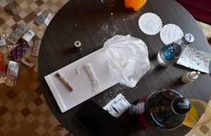 Na zdjęciu na stoliku biały proszek, zwinięty w rulon banknot o nominale 500 złotych, napój w plastikowej butelce,
Na podłodze opakowania różnych tabletek.