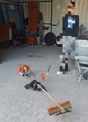 na zdjęciu na ziemi lezy kosa spalinowa, dalej zamazana postać. W tle krzesło.