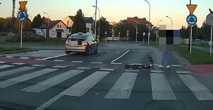 Na zdjeciu przejście dla pieszych. Na lewym pasie, za przejściem dla pieszych pojazd. Na prawym pasie na przejściu leży hulajnoga, obok stoi mężczyzna.