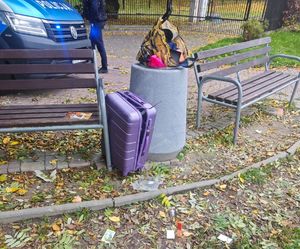 Na zdjęciu w betonowym koszu na śmieci walizka koloru fioletowego, za koszem policyjny radiowóz i umundurowany policjant.