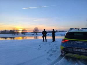 Na zdjęciu  dwóch umundurowanych  policjantów kontroluje teren zbiornika wodnego. Obok radiowóz.