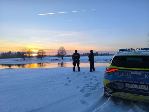 Na zdjęciu  dwóch umundurowanych  policjantów kontroluje teren zbiornika wodnego. Obok radiowóz.