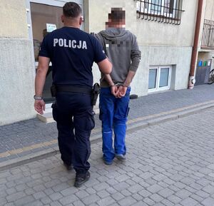 Na zdjęciu umundurowany policjant prowadzi mężczyznę, który na ręce trzymane z tyłu ma założone kajdanki.