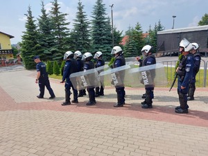 Umundurowanie policjanci idą w dwóch szeregu za dowódcą drużyny. Kilku funkcjonariuszy trzyma w ręku tarcze.