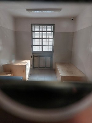 wnętrze pomieszczenia dla osób zatrzymanych ze stolikiem i łóżkami, w środkowej części okno