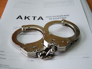 Na zdjęciu znajdują się kajdanki oraz biała teczka z napisem "AKTA".