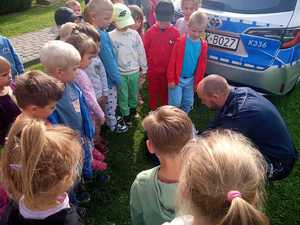 Na pierwszym planie zdjęcia widać grupę dzieci, które stoją wokoło policjanta asp. Tomasza Hałki, który kuca i prezentuje dzieciom jak wygląda wnętrze torby medycznej jaką na wyposażeniu mają policjanci w radiowozie. Po prawej stronie widać trzymaną w ręku tarczę do zatrzymywania pojazdów. Za grupą dzieci i policjantem widać tył radiowozu oznakowanego z otwartymi przednimi drzwiami i po jego lewej stronie również widać grupę dzieci.