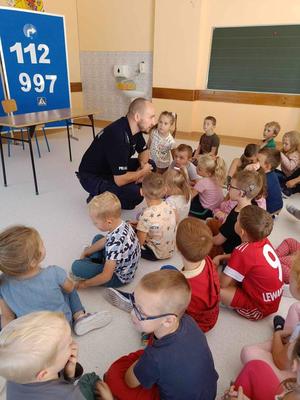 Na zdjęciu w jego środkowej części widoczny jest umundurowany asp. Tomasz Hałka, który kuca przed grupą dzieci z przedszkola w Domaradzu, którzy siedzą na podłodze. Za plecami Policjanta widać stojącą w poprzek ławkę, a za nią tablicę koloru błękitnego z na której od góry widać znak &quot;nakaz skrętu w prawo&quot;, a pod tym znakiem numery alarmowe 112 oraz 997, poniżej widoczny znak przejścia dla pieszych. dalej po prawej stronie widać miejsce w klasie szkolnej przeznaczone na umywalkę, obłożone tarakotą a po prawej stronie widoczna tablica szkolna.