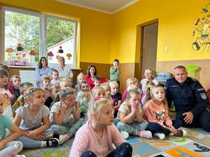 Zdjęcie przedstawia patrząc od prawej część sali przedszkolnej, gdzie odbywa się spotkanie z dziećmi i policjantami. Po prawej stronie na krzesełku dziecięcym siedzi chłopiec ubrany w czapkę policyjną furażerkę a w dłoniach trzyma tarczę do zatrzymywania. Dalej od prawej widać pochyloną umundurowaną policjantkę kom. Monikę Hędrzak, która pokazuje tablice edukacyjne i przeprowadza z dziećmi rozmowę o bezpieczeństwie. W tle za nią widać uśmiechniętego opiekuna mężczyznę, który stoi w progu drzwi. dalej po lewej strony od dołu zdjęcia widać dzieci siedzące na dywanie w sali i patrząc w górę zdjęcia widać siedzącego pośród dzieci umundurowanego policjanta kom. Dariusza Hędrzaka a za nim po lewej Dyrektor przedszkola i zarazem opiekunkę dzieci.