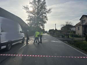 Na zdjęciu od lewej strony kadru widoczny stojący samochód ciężarowy typu bus. Obok niego po prawej stronie widoczny świadek zdarzenia oraz policjanci wydziału kryminalnego ubrani w odblaskowe kamizelki odblaskowe pracujący na miejscu zdarzenia. Na środku kadru widać drogę powiatową z nawierzchnią asfaltową. Po prawej stronie na mostku prowadzącym do posesji widać troje strażaków ubranych w mundury. Z prawej strony kadru widać ciąg zabudowań jednorodzinnych.