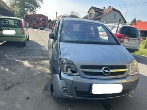 Na zdjęciu w środkowym kadrze na pierwszym planie widoczny samochód osobowy marki Opel, który brał udział w zdarzeniu drogowym. Na pojeździe z jego prawej strony, a patrząc na zdjęcie po prawej stronie widać uszkodzenia przedniego reflektora prawego oraz wgięcie zderzaka po prawej stronie i wgięcia błotnika po prawej oraz rozbita szyba czoło po prawej stronie pojazdu. dalej po lewej stronie iwdać zaparkowany pojazd osobowy marki fiat. po prawej stronie z boku widać tył bozu bojowego straży pożarnej. na środku z tyłu widać kawałek taśmy oddzielającej miejsce zdarzenia. dalej widać bokiem stojący wóz bojowy straży pożarnej. Po prawej stronie kadru widoczne zabudowania jednorodzinne.