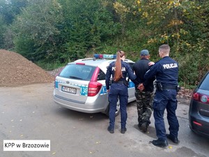 Na zdjęciu od lewej strony widać usypaną hałdę ziemi. Dalej na lewo widać stojący policyjny oznakowany radiowóz. Przed radiowozem od lewej policjantka w umundurowaniu trzymająca za lewe ramię zatrzymanego zakutego w kajdanki mężczyznę a po prawej stronie trzymający za prawe ramię umundurowanego policjanta. Dalej na prawo widać lewy tył innego pojazdu. Na tle zdjęcia widoczne zadrzewienia.