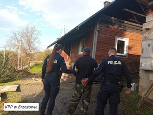 Na zdjęciu w dolnym lewym rogu na biały tle napis KPP w Brzozowie. Na środku kadru widoczni umundurowani policjanci z lewej policjantka z prawej policjant a pomiędzy nimi na środku zatrzymany mężczyzna w kajdankach sprawca dokonania podpalenia. Na środku kadru w na drugim tle widać strawiony ogniem dom jednorodzinny. Za tym domem nad dachem widoczny szczyt wieży kościelnej. Po prawej stronie kadru widoczna część murowana budynku na której ścianie widać dwie skrzynki wysokiego napięcia. Po lewej stronie kadru na ostatnim tle widać zadrzewienia i infrastrukturę energetyczną w postaci linii i słupów energetycznych.