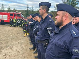 Zdjęcie przedstawia od prawej strony ustawionych w rzędzie biorących udział w ćwiczeniach. W pierwszej kolejności są to umundurowani policjanci, a dalej w tle umundurowani druhowie OSP, za którymi widać strażacki wóz bojowy. Tło zdjęcia to zadrzewienia leśne i niebo.