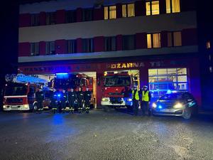 Zdjęcie przedstawia zgromadzonych na placu Komendy Powiatowej Państwowej Straży Pożarnej w Brzozowie umundurowanych funkcjonariuszy policji i straży wraz z strażackimi wozami bojowymi i oznakowanym radiowozem policyjnym. Pojazdy służbowe mają włączone sygnały świetlne barwy niebieskiej. Z tyłu za nimi budynek Komendy Powiatowej Państwowej Straży Pożarnej w Brzozowie.