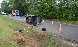 Uszkodzony pojazd leży na boku na jezdni. W tle służby ratunkowe.