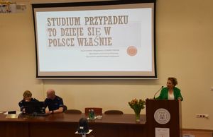 Prelegentka przy mównicy. Obok policjant i policjantka. W tle wyświetlony jest slajd prezentacji.