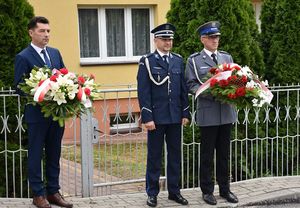 Mężczyzna w garniturze z wiązanką kwiatów i dwóch policjantów w mundurach galowych. Jeden z nich trzyma wiązankę kwiatów