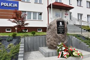 Tablica upamiętniająca, pod nią wiązanki kwiatów. W tle budynek Komisariatu Policji w Pruchniku.