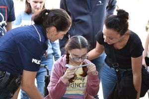 Policjantka stoi przy dziewczynce. Dziewczynka czyta. Obok niej znajduje się też kobieta.