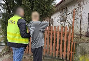 Policjant w kamizelce trzyma za rękę zatrzymanego mężczyznę. W tle budynek mieszkalny