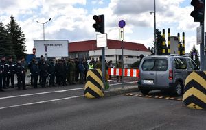 Na drodze widoczny jest samochód. Na chodniku stoją mundurowi oraz osoby cywilne