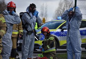 Strażacy podczas zakładania kombinezonu ochronnego. W tle radiwoóz