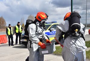 Strażacy w kombinezonach. W tle policjanci