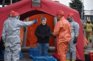 Mężczyzna przed namiotem. Obok niego strażacy w kombinezonach