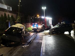 Zdarzenie drogowe. Pora nocna. na jezdni znajdują się dwa uszkodzone pojazdy. W tle wóz strażacki.