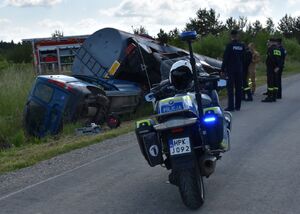 Policjant na motocyklu. W tle służby ratunkowe i przewrócona cysterna oraz samochód
