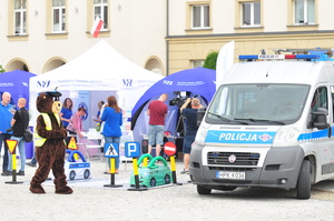 Radiowóz i policyjny miś. W tle stoiska NFZ, dziennikarka, kamerzysta