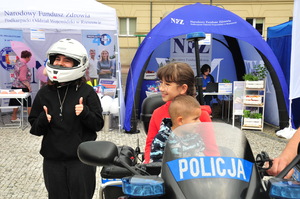 Dziewczynka oraz chłopiec siedzący na policyjnym motocyklu. Obok dziewczyna w kasku unosząca dwa kciuki do góry