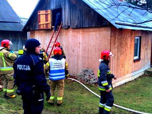 Policjant oraz kilki strażaków przed drewnianym domem