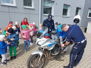 Policjant i policjantka pomagający wsiąść na motocykl chłopczykowi. Obok stoją przedszkolaki