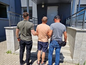 Nieumundurowani policjanci, którzy stoją po obu bokach, trzymając za ręce zatrzymanego mężczyznę, przed budynkiem komendy