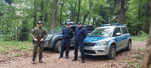 Policjant oraz strażnicy na tle lasu i pojazdów służbowych