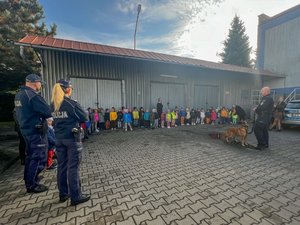 Zdjęcie przedstawia funkcjonariuszy Komendy Powiatowej Policji w Leżajsku. Dwoje z nich stoi po lewej stronie kadru, a z kolei trzeci, przewodnik psa, stroi po prawej stronie kadru. Zwróceni oni są wszyscy w kierunku dzieci, stojących przy garażach, na dziedzińcu Komendy. Na zdjęciu widać psa służbowego podczas pokazów.